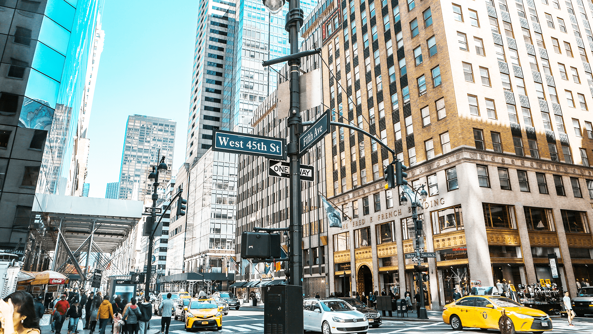 Krydset mellem Fifth Avenue og West 45th Street i New York City med høje bygninger, travle fodgængere og gule taxaer i forgrunden.