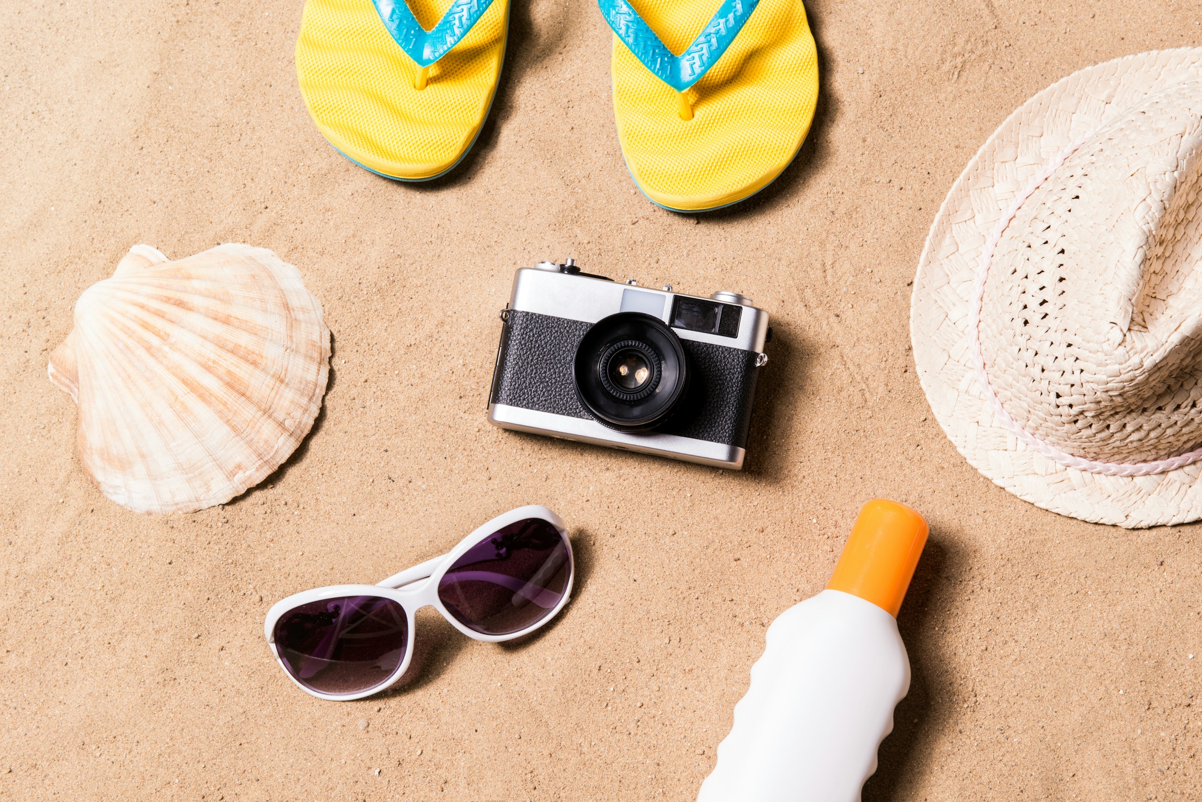 Rejseprodukter såsom en hat, kamera og solcreme på en sandstrand