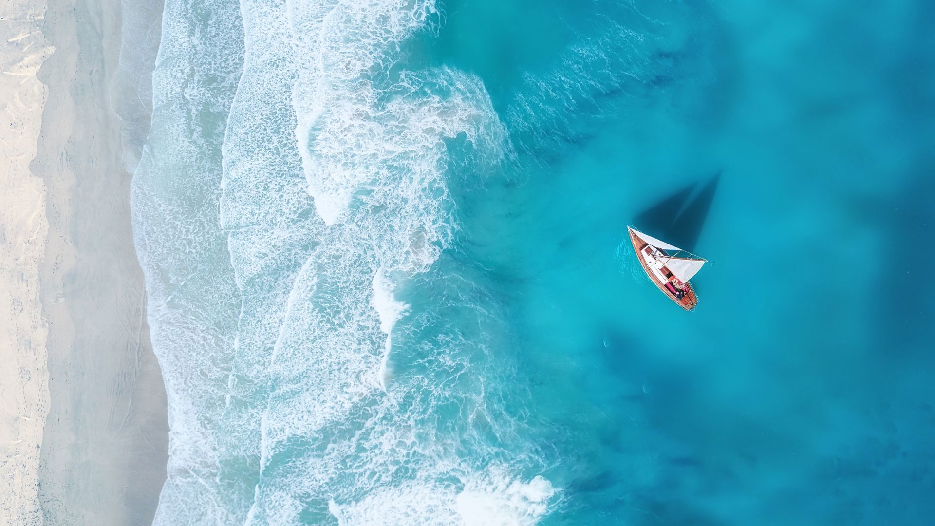 A sailboat crossing the turquoise water near the white foaming waves of the beach, seen from above.