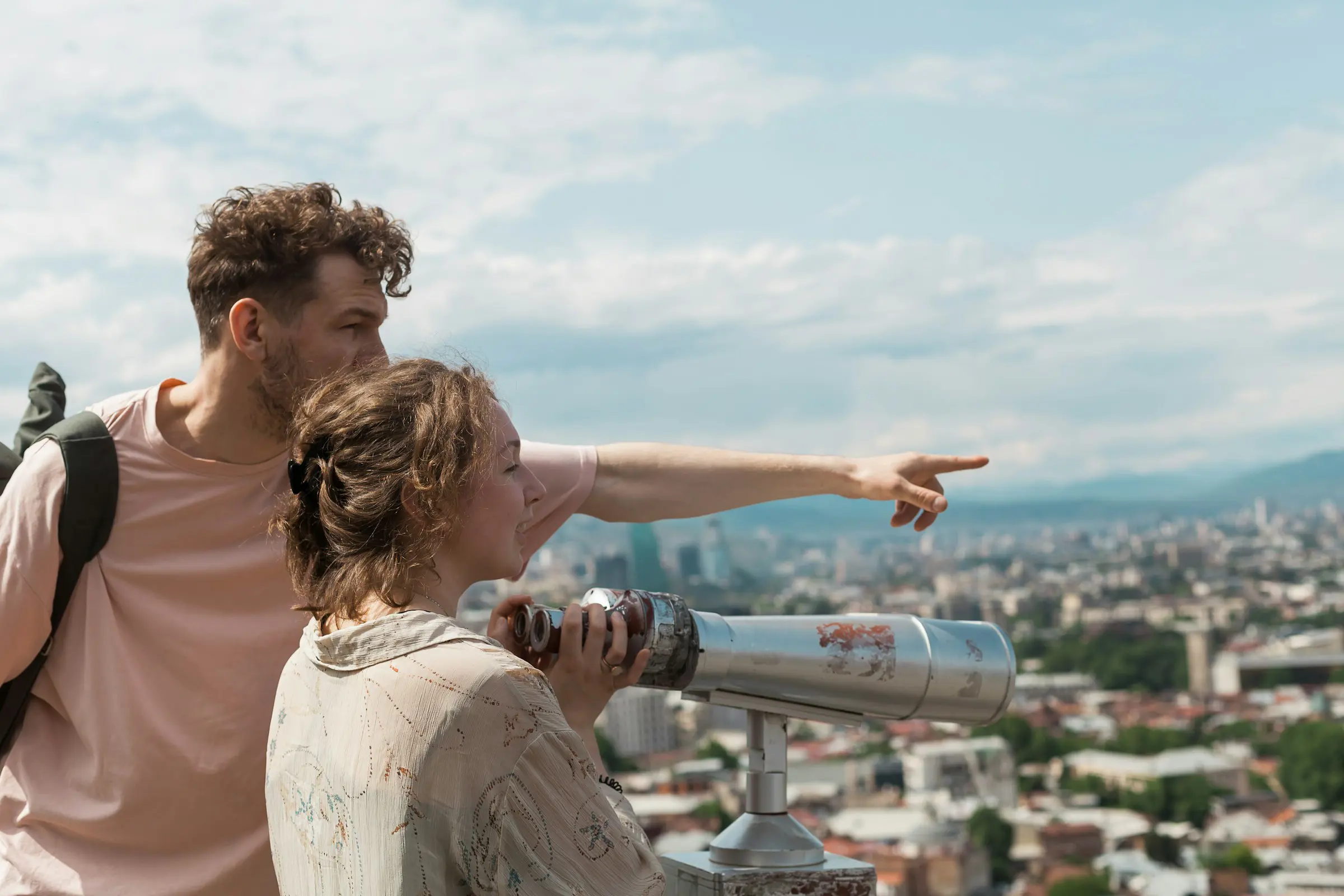 Couple looking at the view of the city from vantage point, man pointing out something in the landscape while woman looks through binoculars.