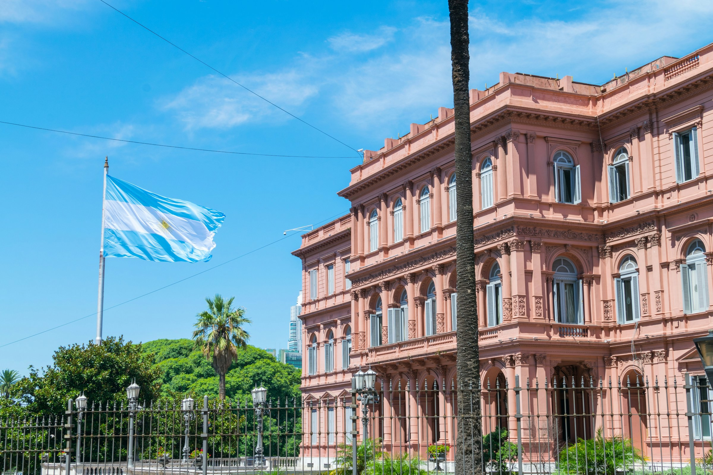 Argentinas flag vajer foran præsidentpaladset Casa Rosada i Buenos Aires under en klar blå himmel med grønne områder i forgrunden.