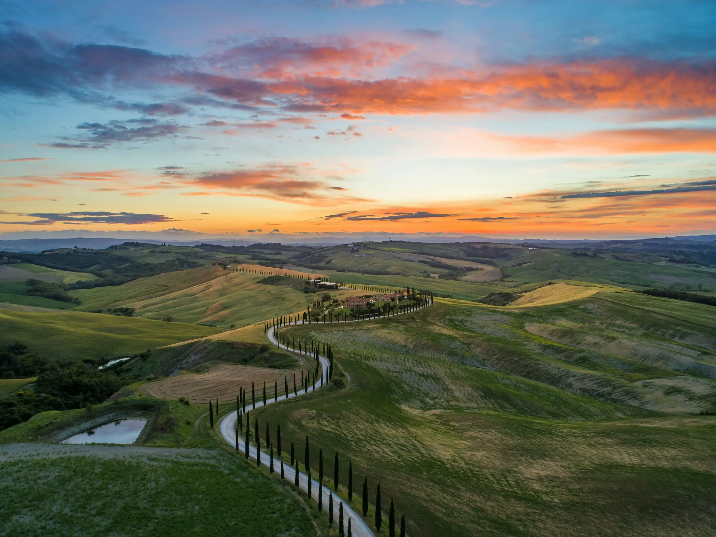 Scenic view of Tuscan countryside at sunset with rolling hills, a winding road lined by cypress trees, and a vibrant sky.