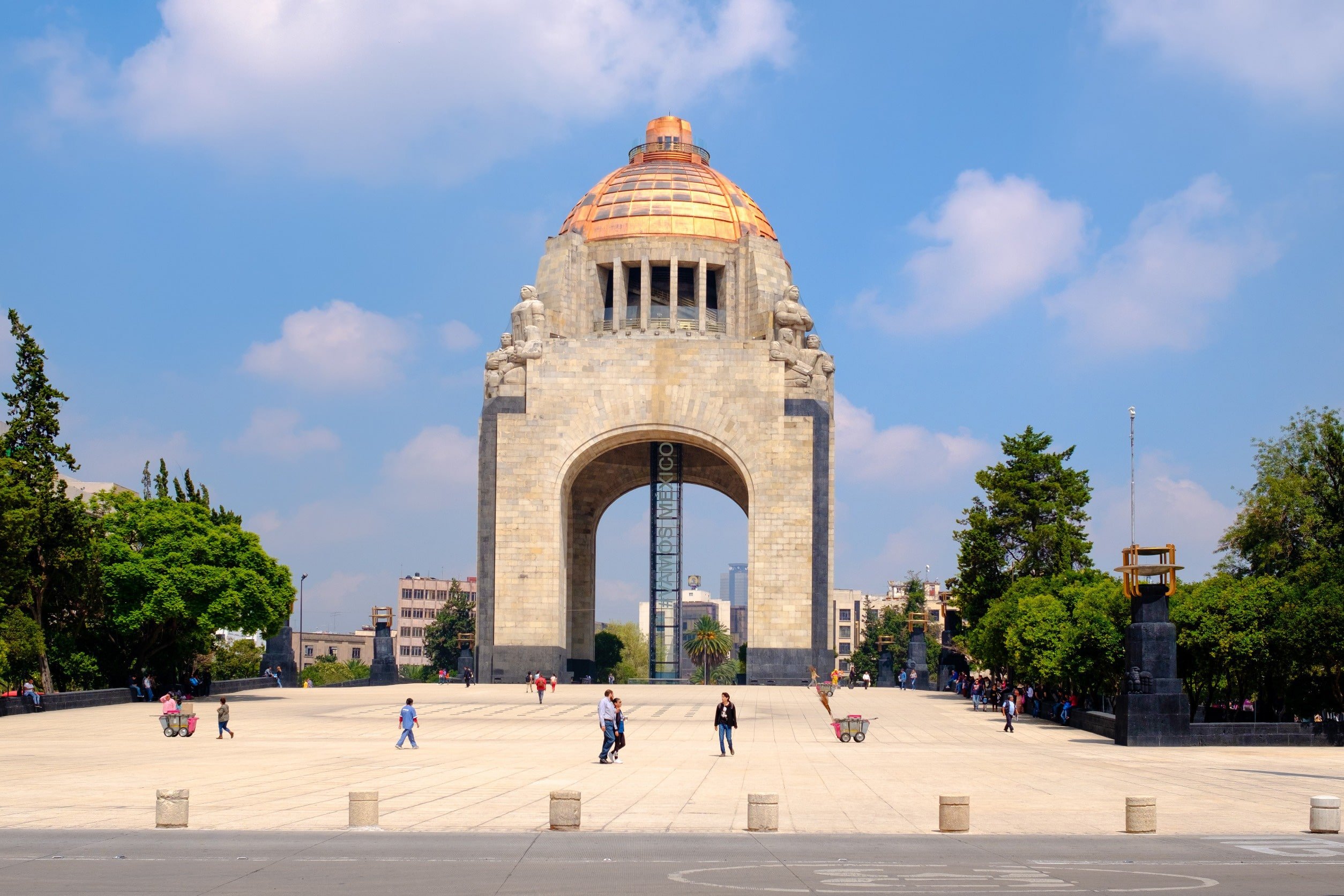 En plads, der fører op til monumenter i Mexico City med grønne træer omkring og blå himmel i baggrunden