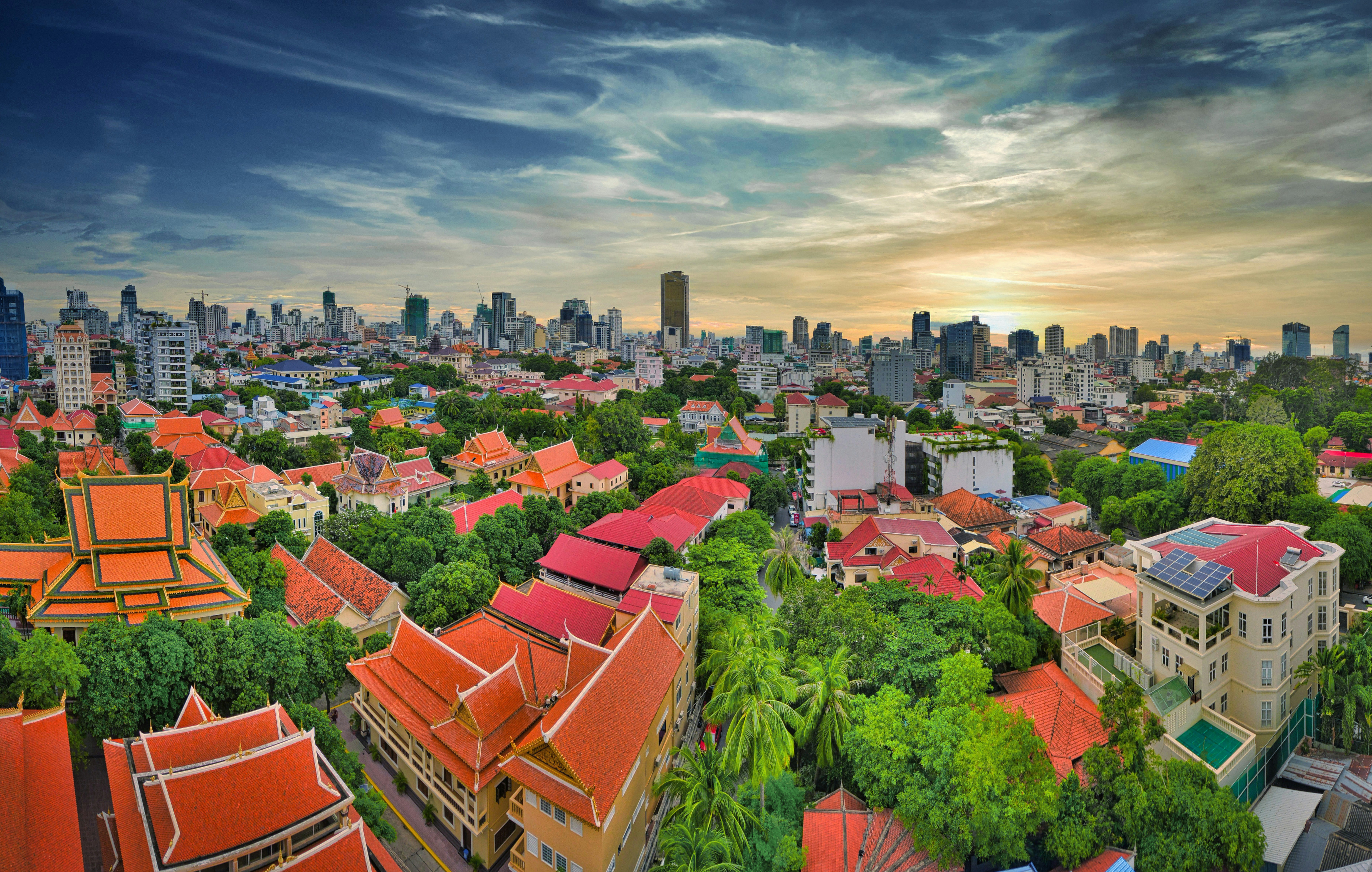 Rejs til Phnom Penh - Panoramaudsigt over Phnom Penh ved solnedgang med traditionelle og moderne bygninger omgivet af frodige træer.