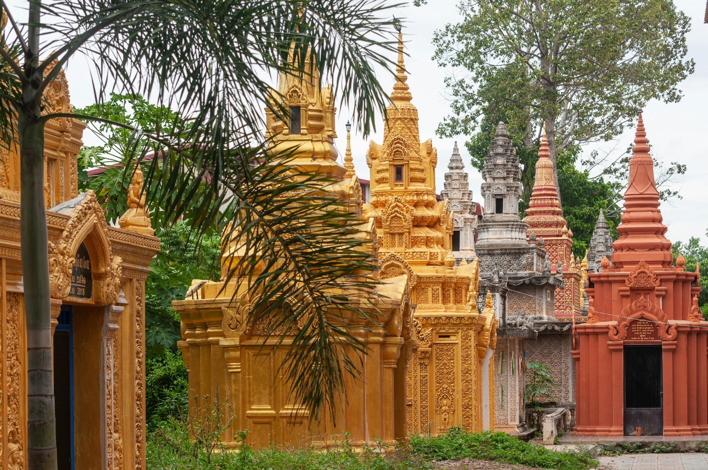 Traditionelle gyldne og terrakotta-stupaer ved et tempel omgivet af grønne områder i Phnom Penh.