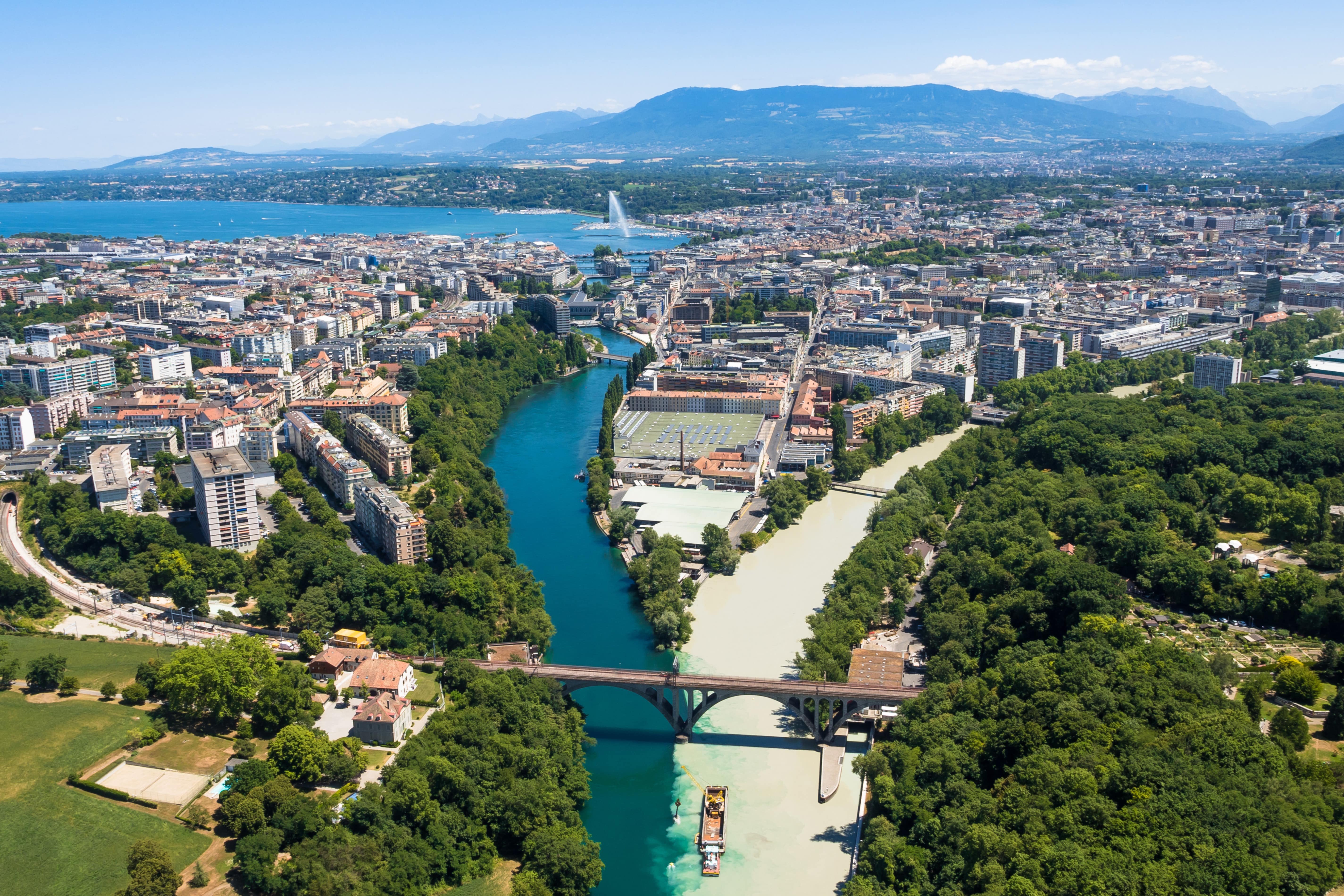 Rejs til Genève - Udsigt fra oven over Genève by med grønne områder og en blå flod, der løber gennem byen og bjergene i baggrunden