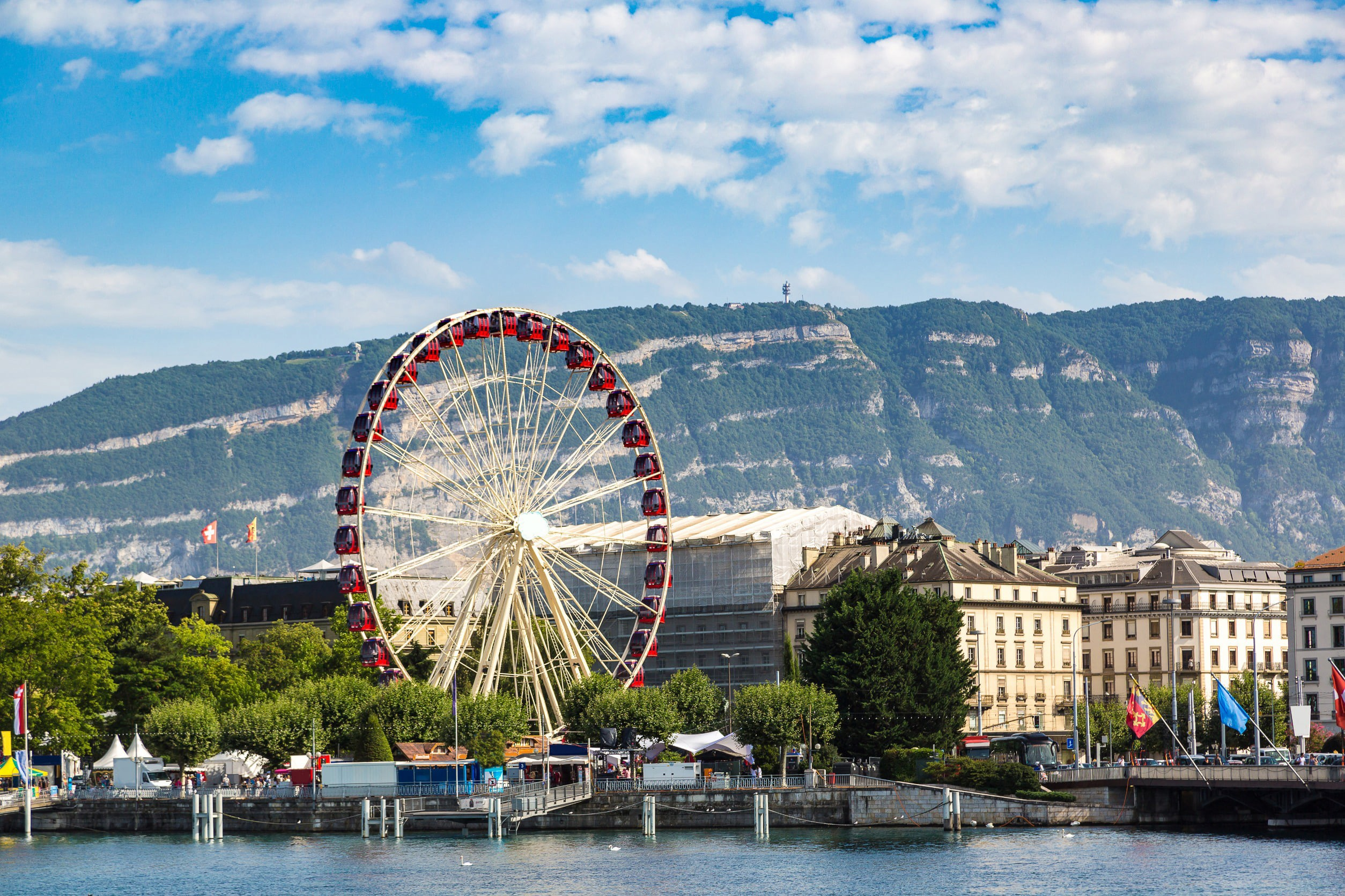 Udsigt over Genève by med bygninger og et pariserhjul og grønne områder omkring den med smukke bakker og blå himmel i baggrunden