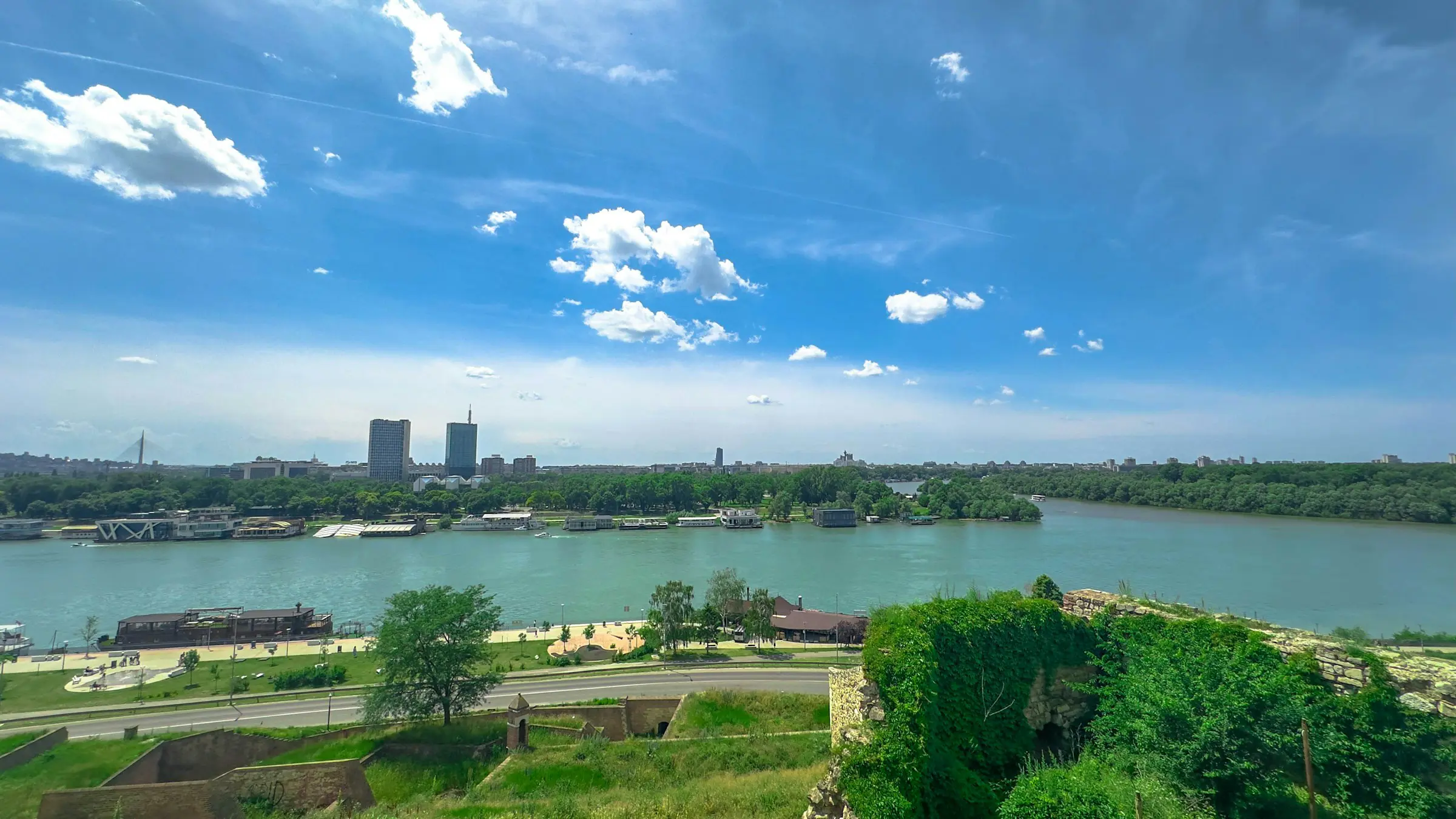 View of a river in a cityscape under a bright blue sky with scattered clouds, featuring green trees along the riverbank and urban buildings in the distance.