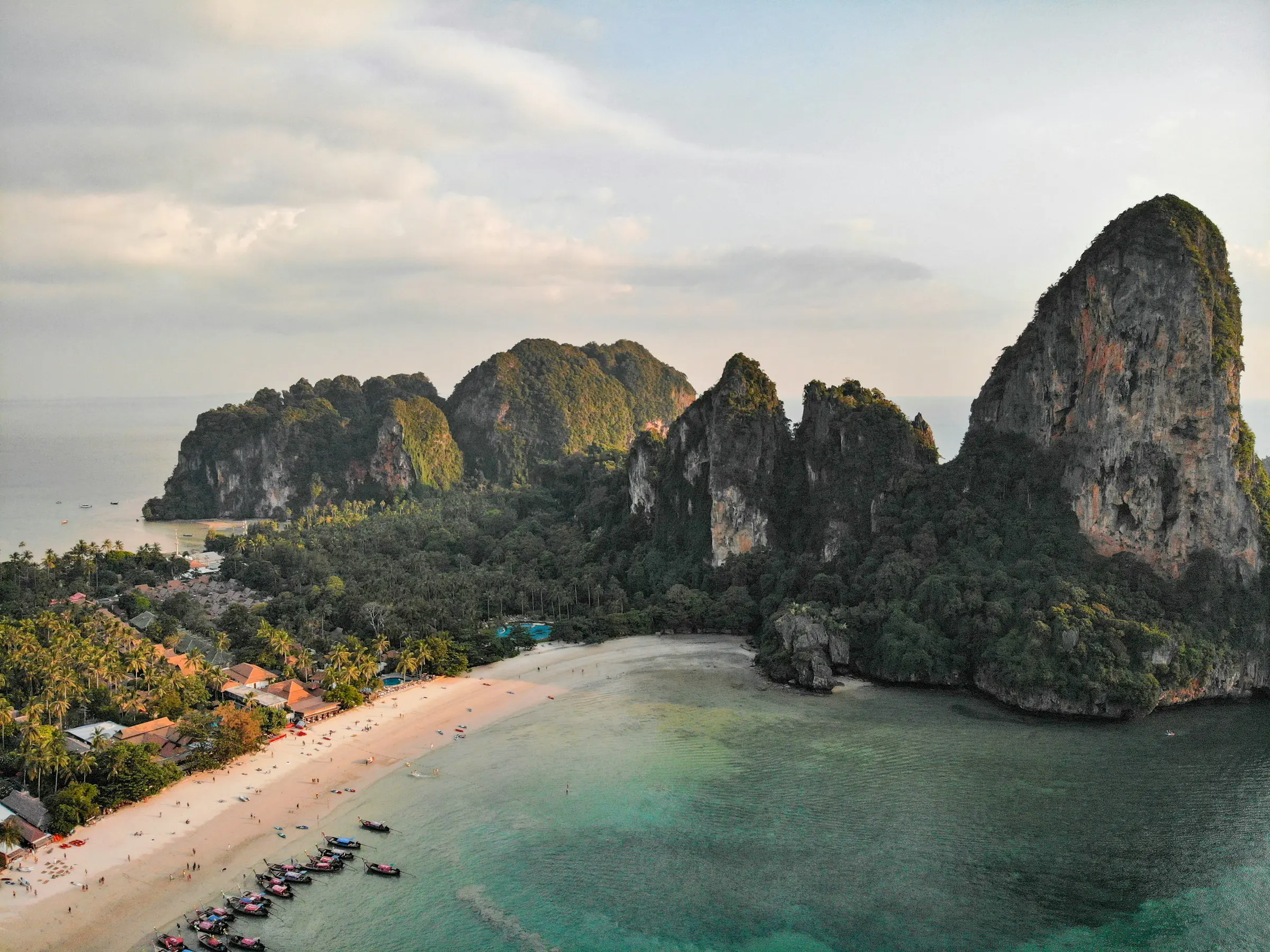 Billedet viser en malerisk strand med hvide sandstrande omgivet af frodige grønne bjerge og klipper, beliggende ved det klare, turkisblå hav i Krabi, Thailand. Klimaet er solrigt med få skyer.